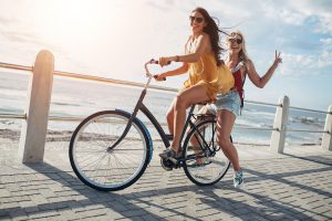 two women riding bikes on a boardwalk near the ocean.