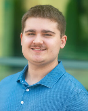 Close-up portrait of a smiling man wearing a blue polo shirt outdoors with a green blurred background.